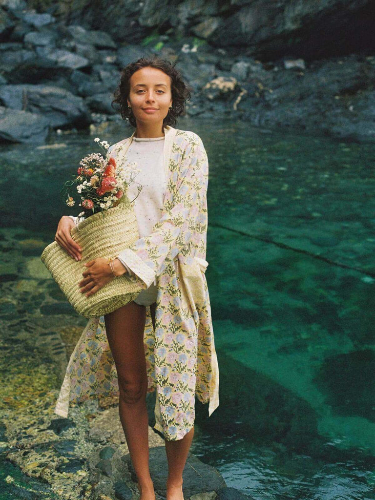 Model by the sea wearing a floral block print robe in buttermilk yellow holding a Bohemia basket filled with dried flowers.