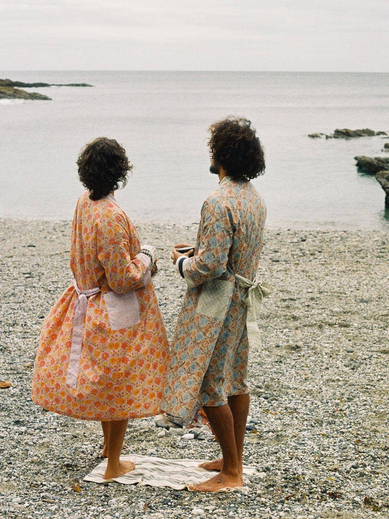 Two models on a pebble beach wearing floral block print robes in duck egg and lilac.