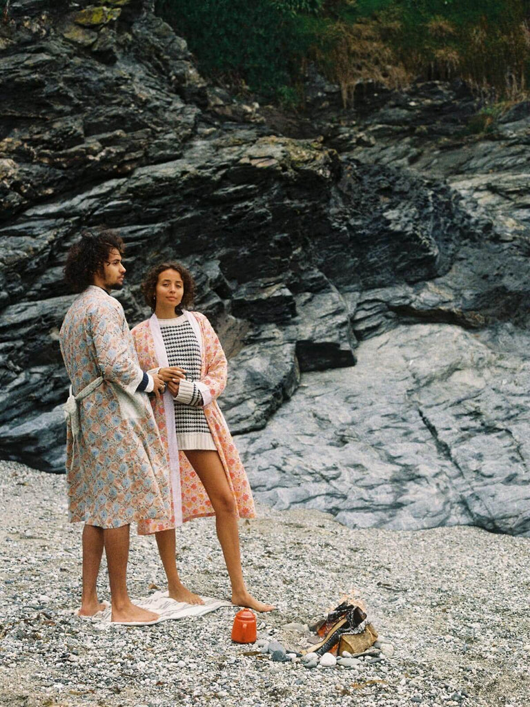 Two models on a pebble beach with camp fire wearing floral block print robes in duck egg and lilac.