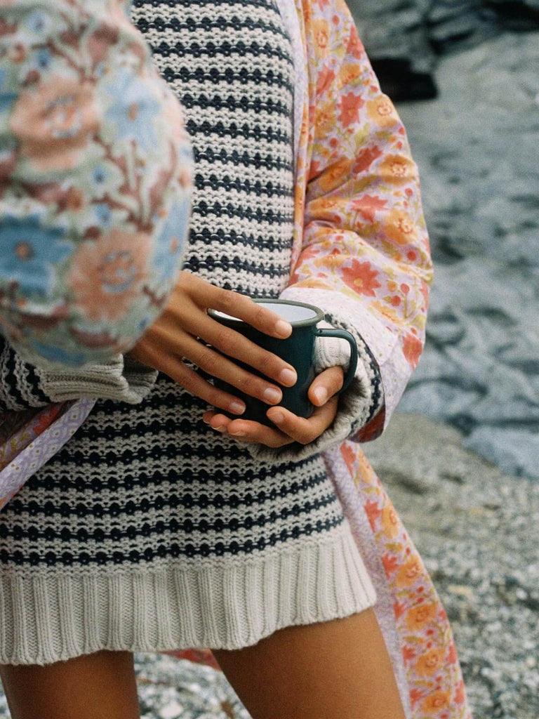 A model wearing a cosy knit and floral block print robe on a pebble beach holding an enamel mug 