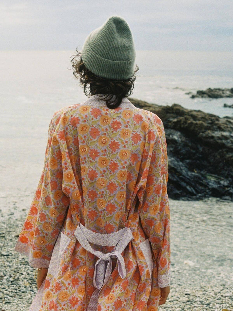 A model wearing a lilac floral block print robe and wool hat on a pebble beach