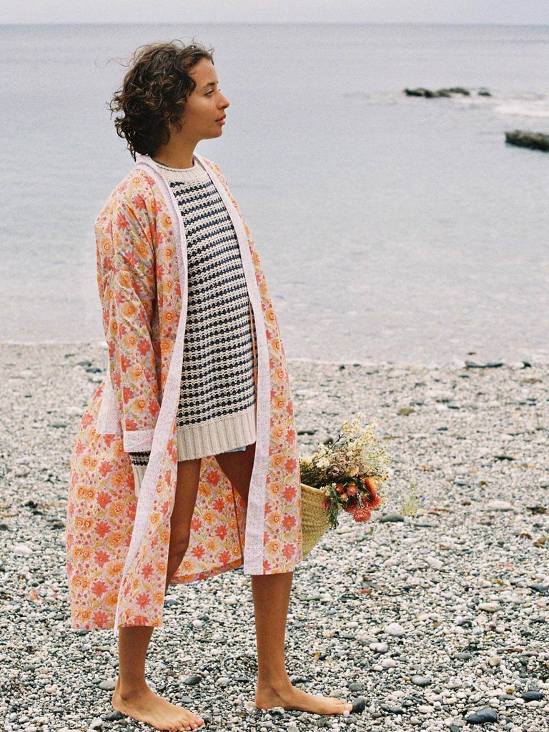 Model on a pebble beach wearing a block print robe in lilac holding a Bohemia basket with dried flowers.