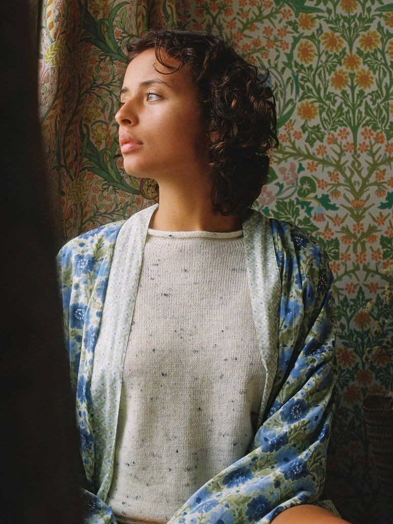 Model sat at a window wearing a sage and blue floral block print robe  with vintage floral wallpaper in the background