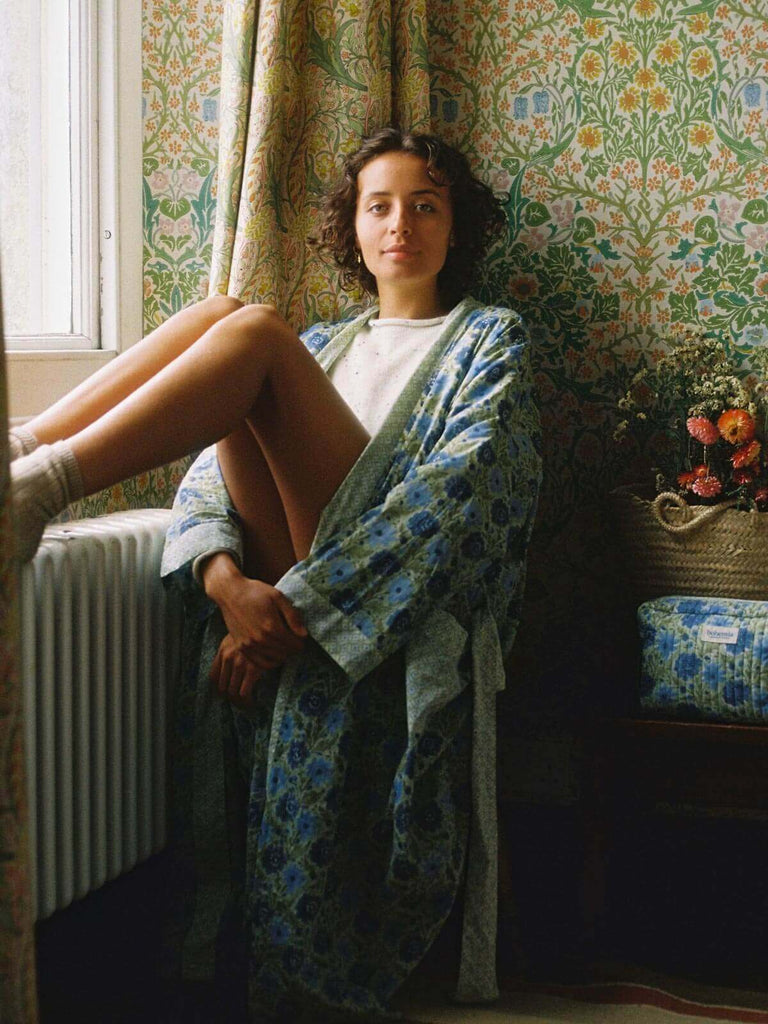 Model sitting by a window in a country house wearing a sage block print robe and quilted house slippers