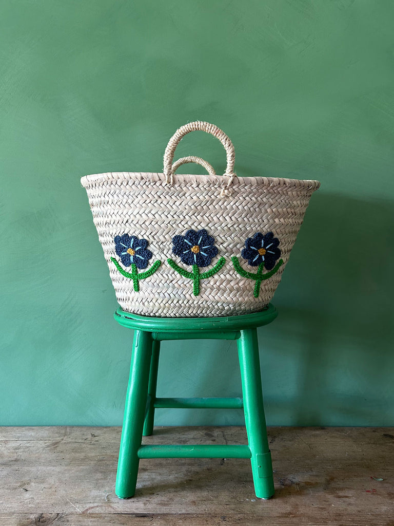 Natural basket with an embroidered daisy motif, resting on a green stool against a vibrant green wall| Bohemia Design