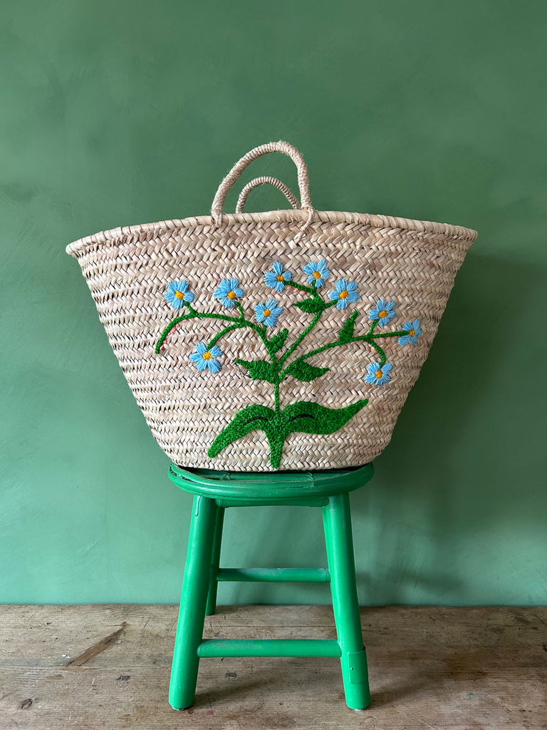 Handwoven market basket with an embroidered Forget-Me-Not flower in delicate sky blue on a green stool against a green wall | Bohemia Design