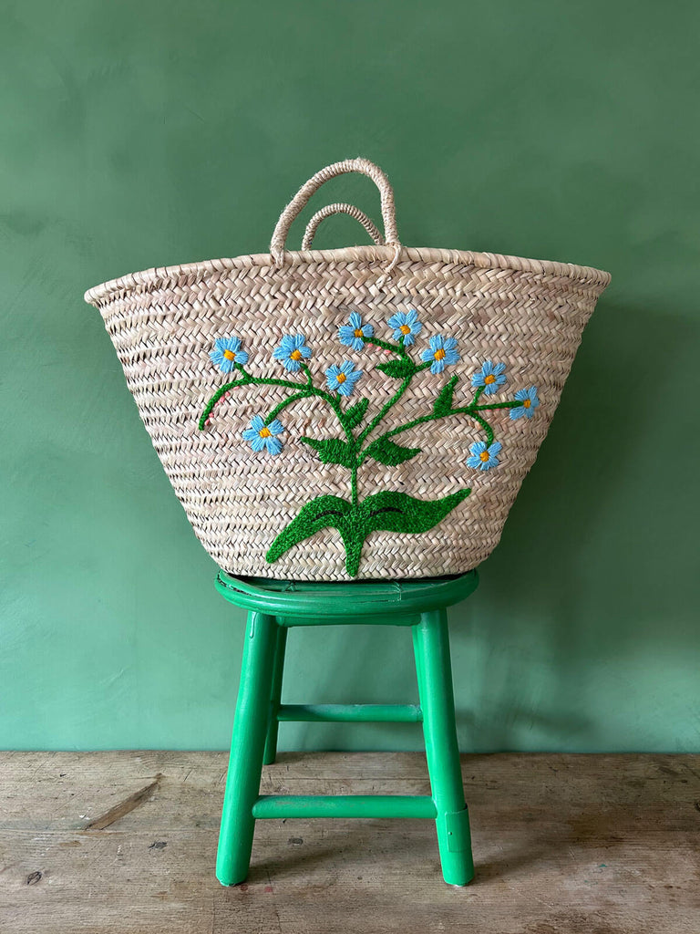 Handwoven market basket with an embroidered Forget-Me-Not flower in delicate sky blue on a green stool against a green wall | Bohemia Design