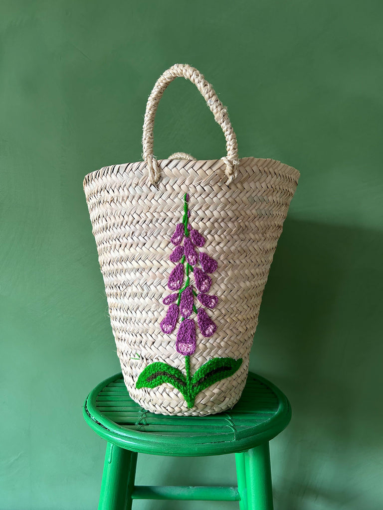 Natural woven basket bag with a hand-embroidered foxglove flower on a green stool against a green wall by Bohemia Design