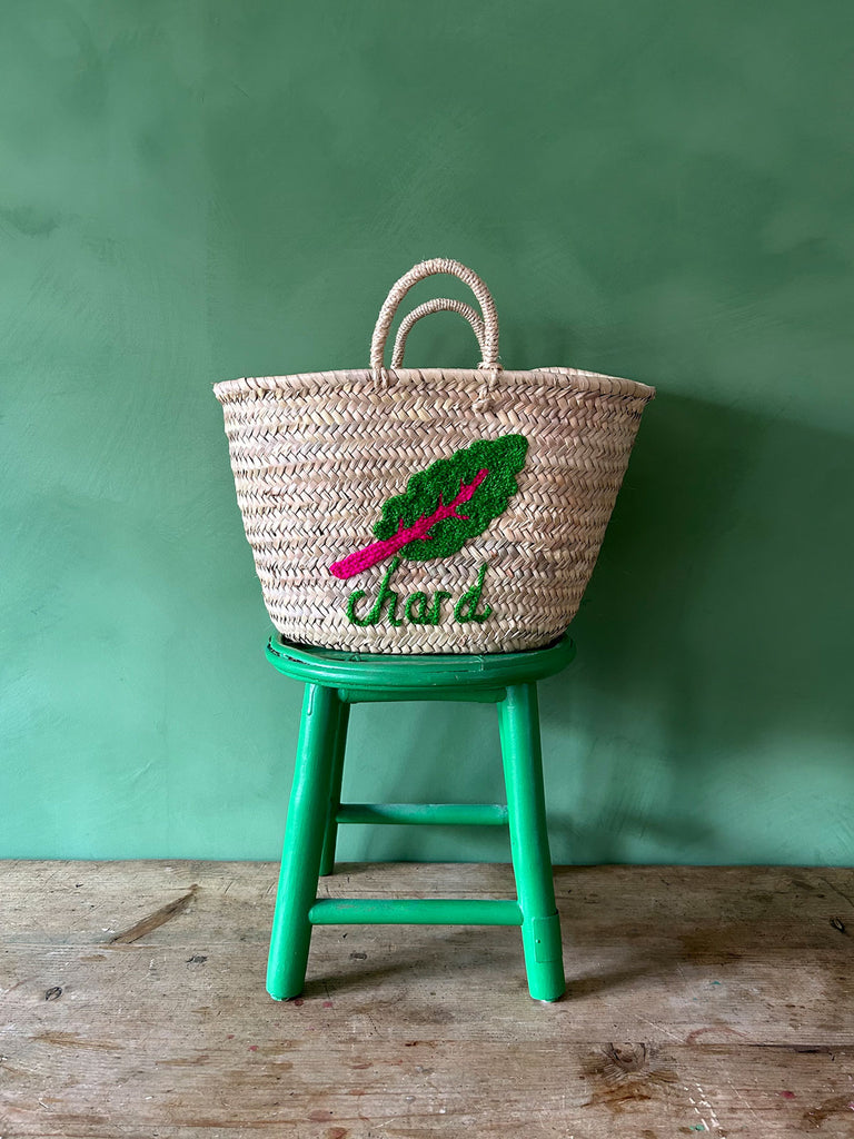 Handwoven palm leaf basket bag with an embroidered chard illustration and typography, resting on a green stool against a green wall | Bohemia Design
