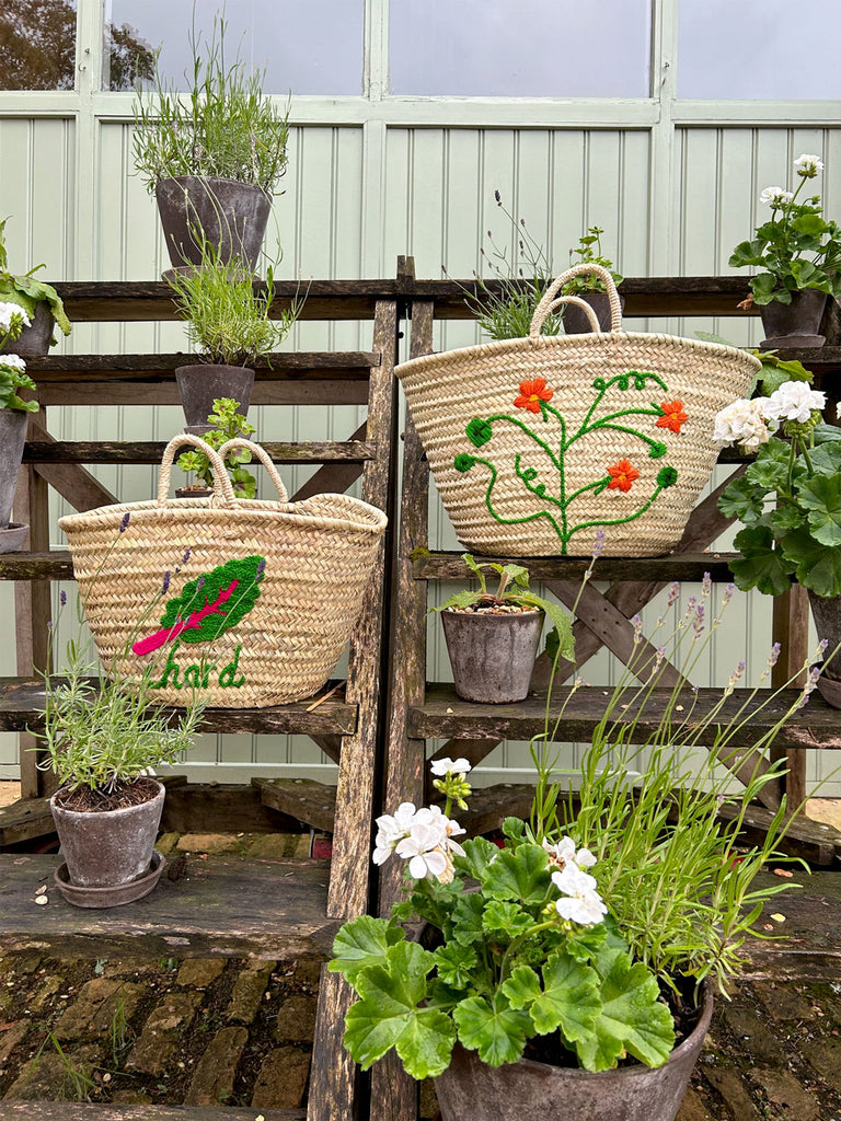 Hand embroidered natural woven basket bags on a rustic wooden display surrounded by garden plants | Bohemia Design