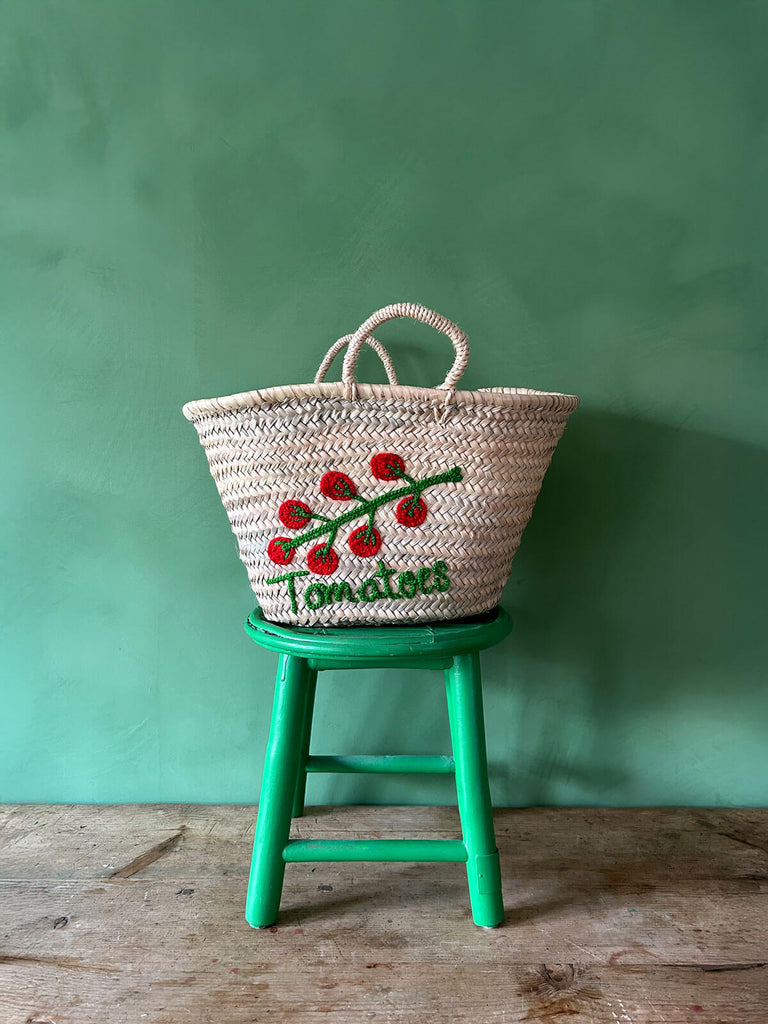 Handwoven wholesale market basket with small handles and an embroidered tomato illustration and accompanying text sitting on a green stool against a green wall | BohemiaDesign