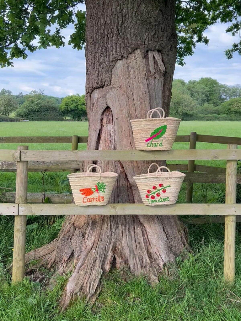 Hand embroidered market baskets featuring three original vegetable designs, all resting on a wooden fence in a picturesque outdoor scene | Bohemia Design