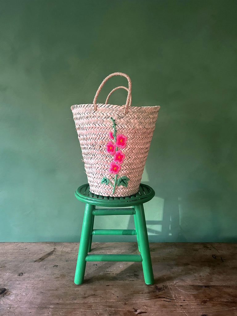 Natural woven basket bag with a hand-embroidered hollyhock flower on a green stool set against a green wall by Bohemia Design