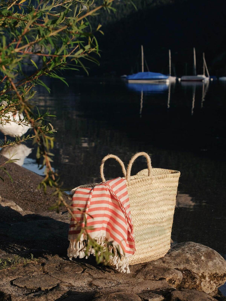 An orange stripe Sorrento hammam towel in a basket on the shore of a river with boats in the distance