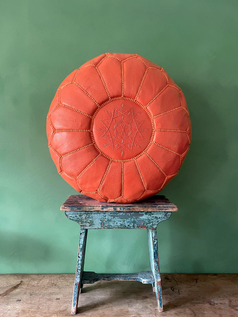 Authentic Moroccan leather pouffe in vibrant orange, placed on a stool against a green wall by Bohemia Design