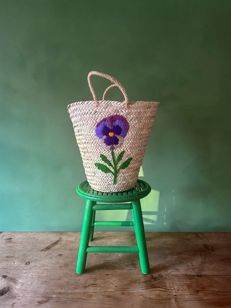 A natural handcrafted basket bag with a hand-embroidered pansy flower displayed on a green stool against a green wall | BohemiaDesign