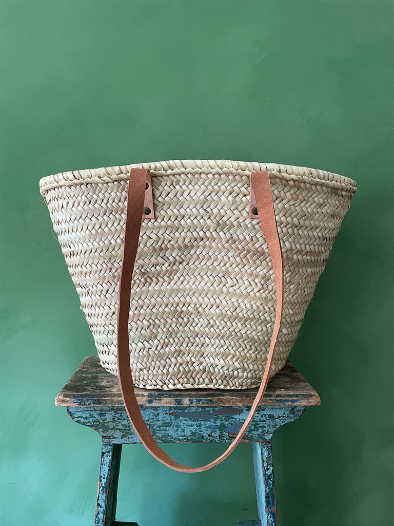 Moroccan shopping basket bag with long natural leather shoulder straps, displayed on a rustic stool against a green wall | BohemiaDesign